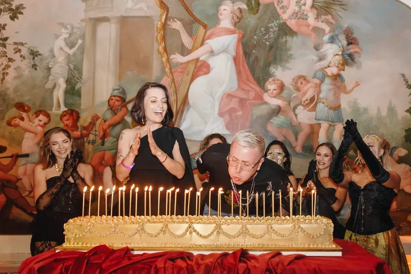 O convidado de honra a apagar as velas do bolo de aniversário de andares dourados diante do fresco do Palácio do Grilo, com produção da BTQ Events