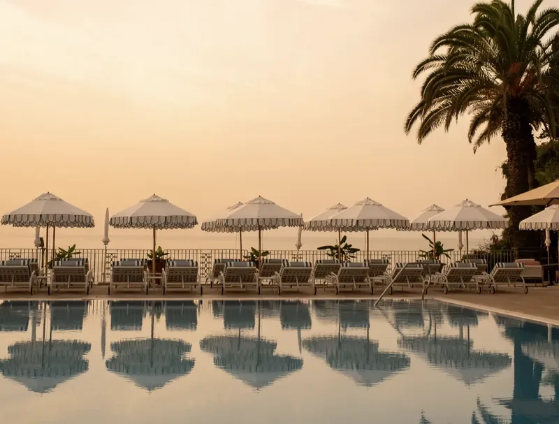 Reid's Palace pool at golden hour · striped umbrellas and palm tree reflected in the water above Funchal Bay